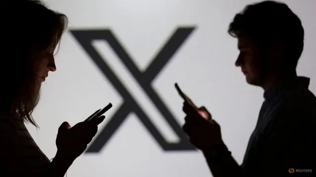 Reuters photo of two people in silhouette looking at their phones in front of a large X logo