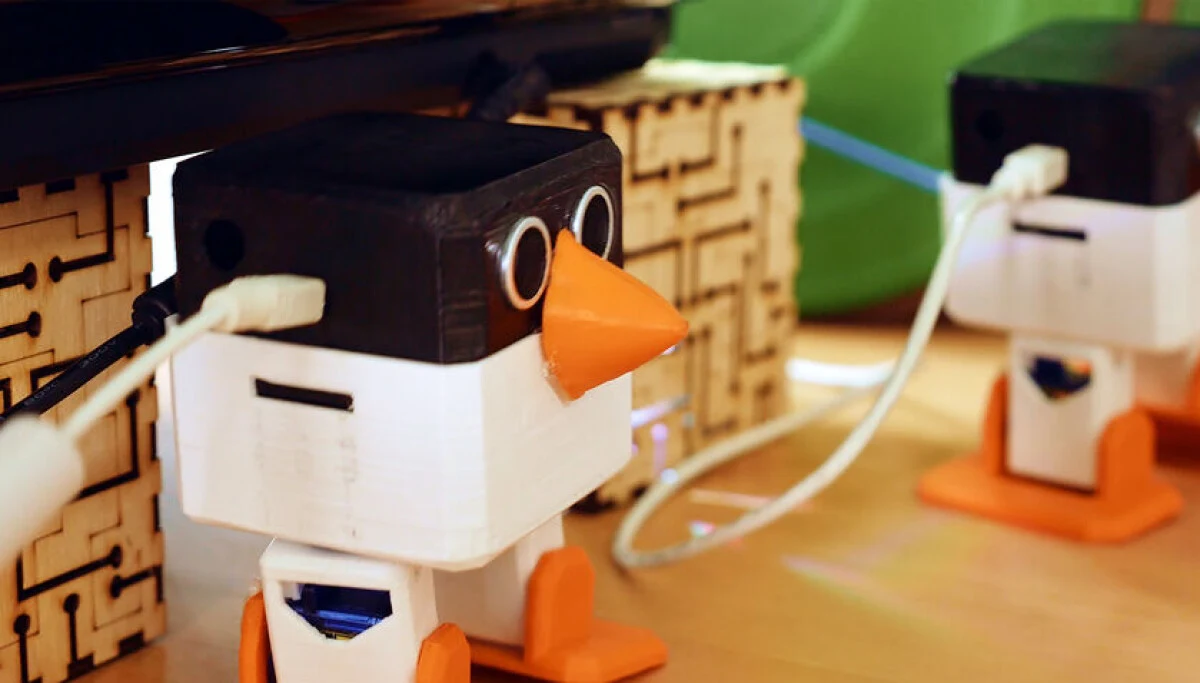 Two 3D-printed penguin-shaped robots with USB cables on a wooden desk.