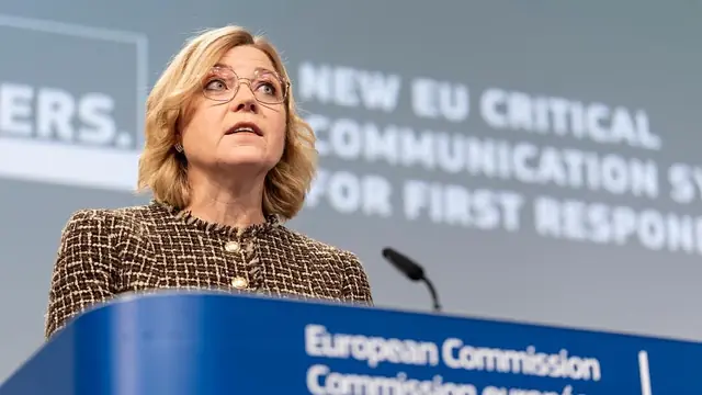 A woman with glasses speaking at a European Commission lectern in front of a blue banner