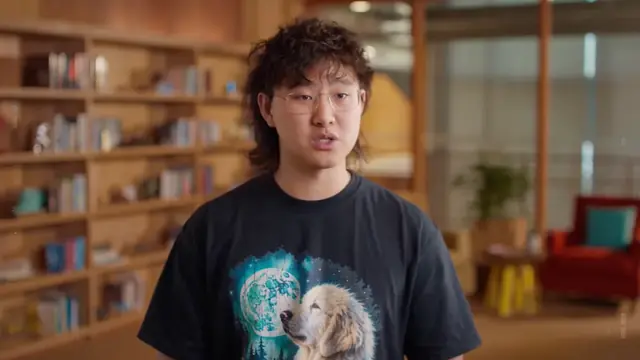 A young man in a dog-print t-shirt speaking in an office with bookshelves in the background