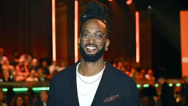 A smiling man in glasses and a dark jacket at an event with stage lights in the background